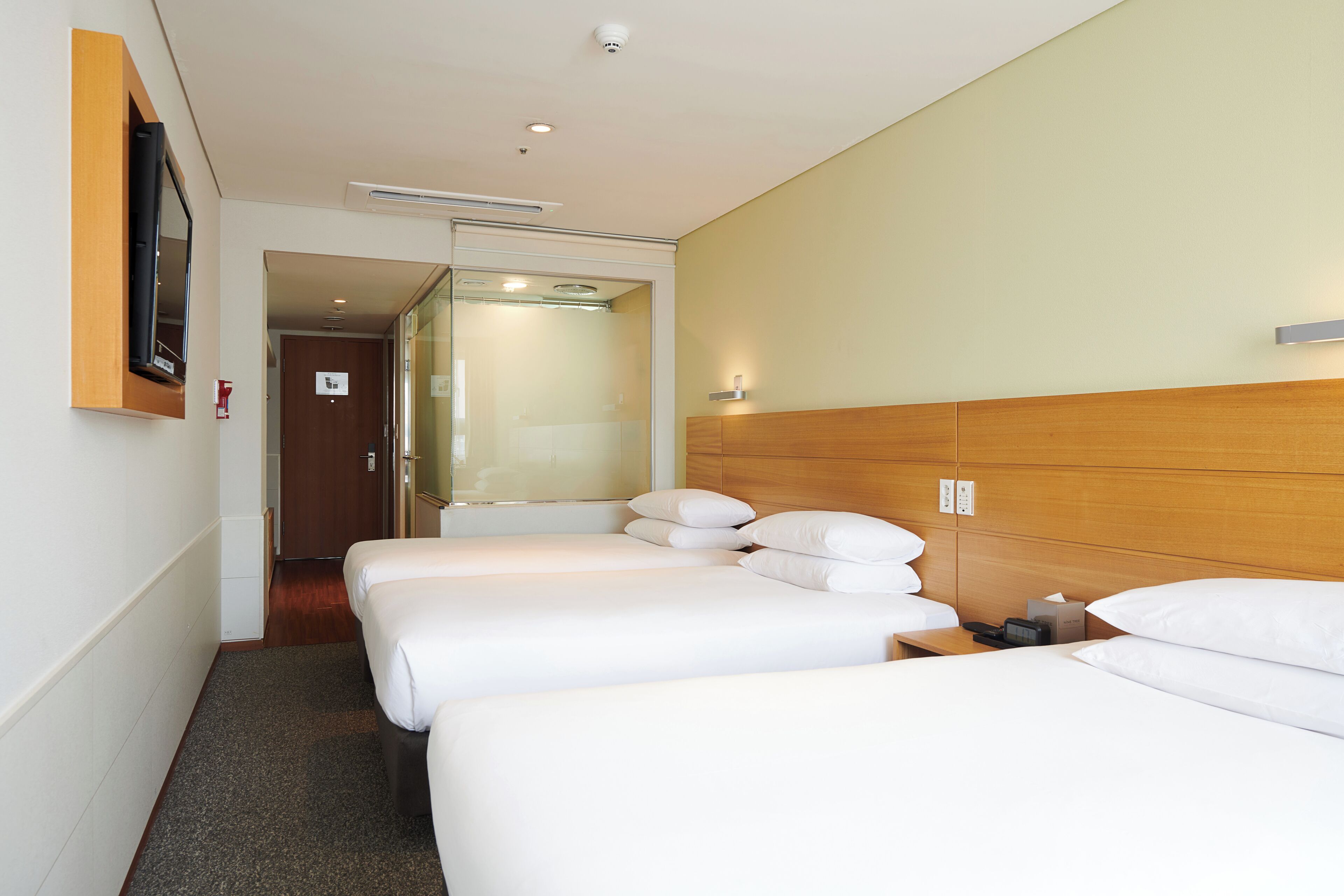 A hotel room with three single beds, a wooden headboard, a bedside table with a lamp, and a flat-screen TV mounted on the wall.