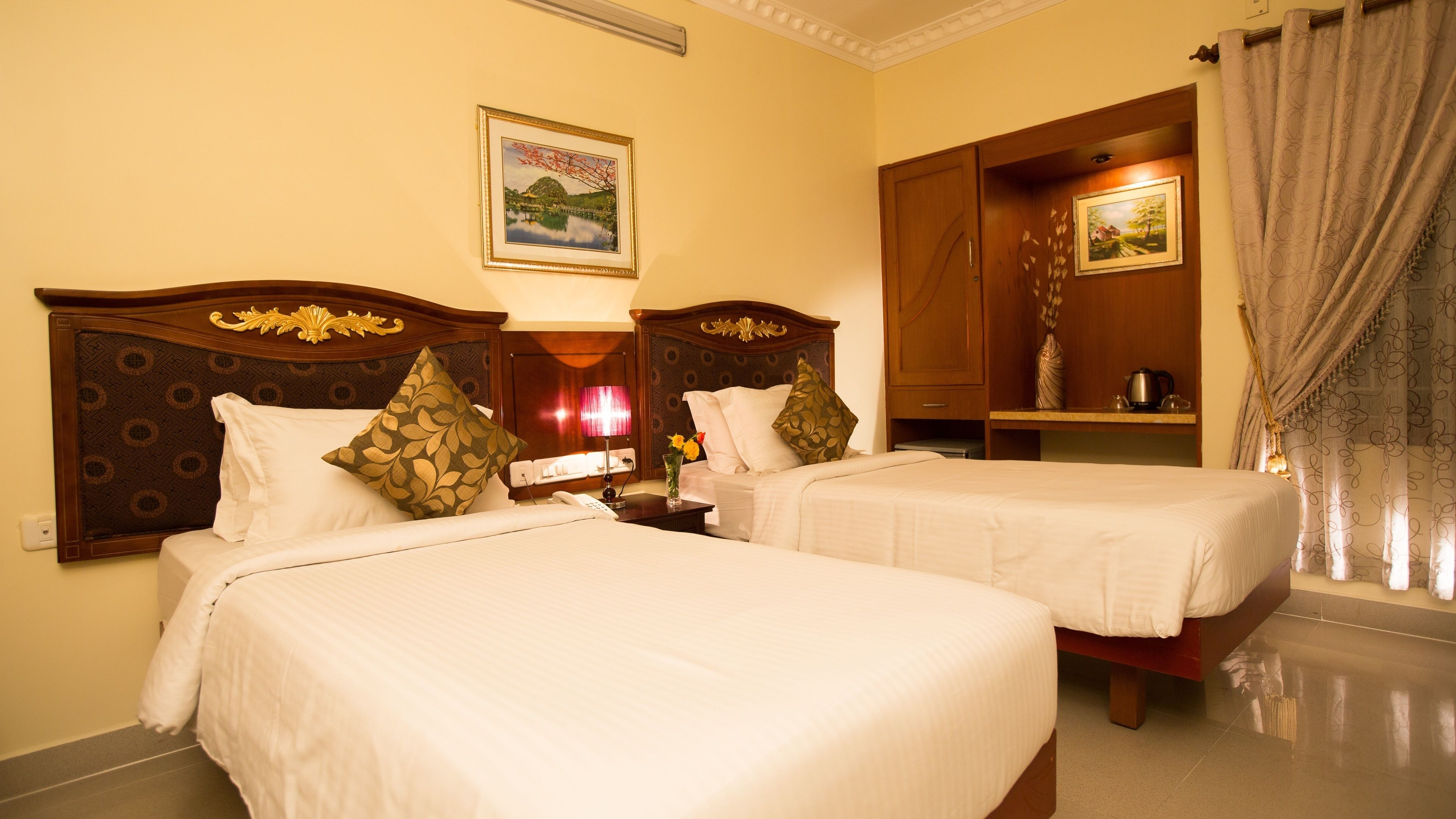 A hotel room with two beds, a nightstand, a lamp, a painting, and a bathroom with a shower curtain.
