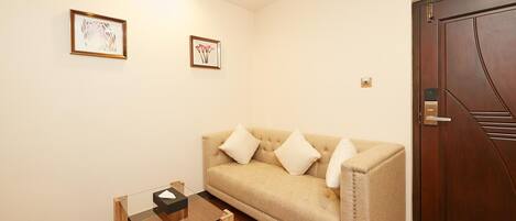 A room with a beige sofa, a glass coffee table, and a wooden floor. There are two framed pictures on the wall and a wooden door.
