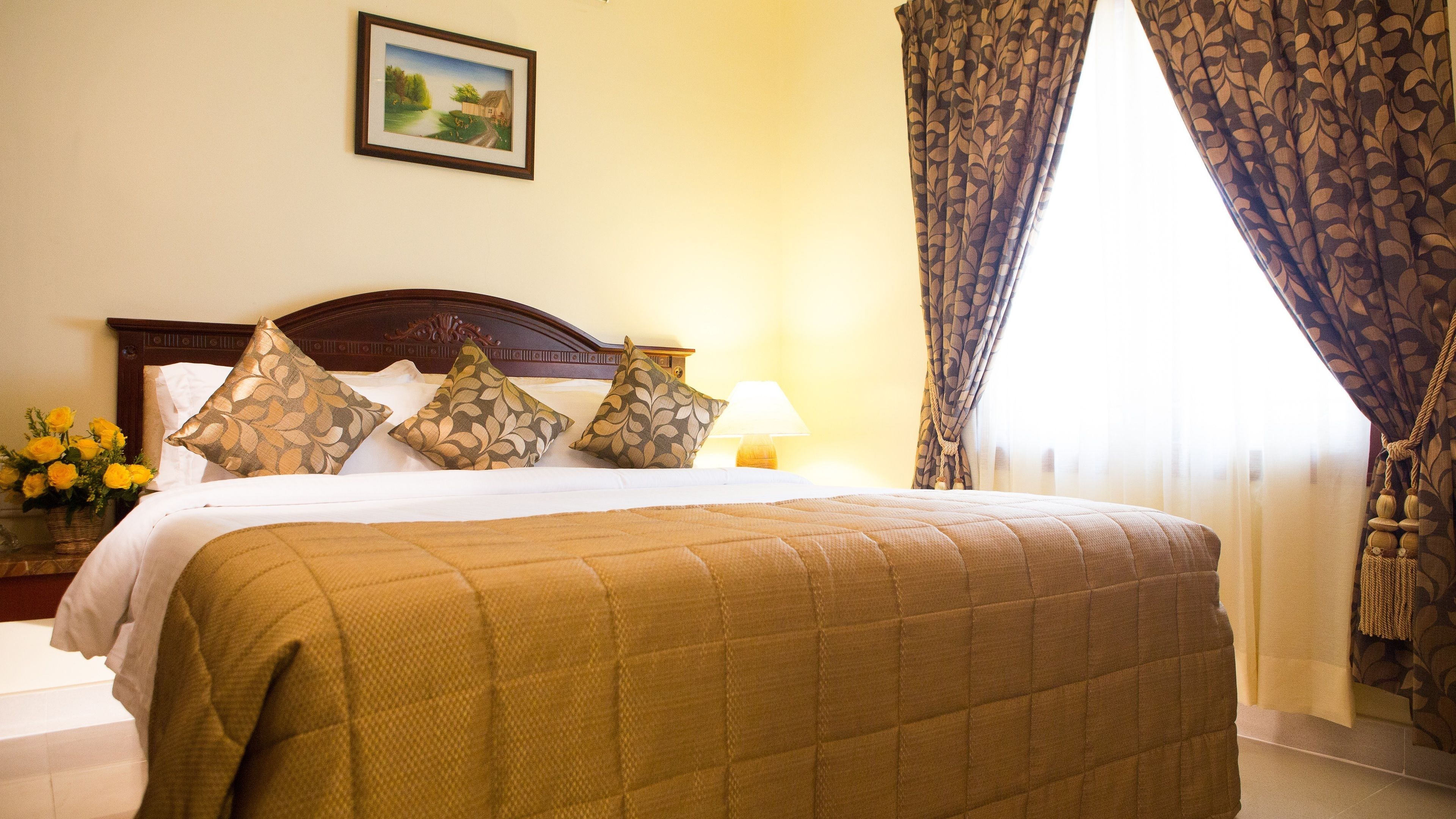 A neatly made bed with a quilted cover, a wooden headboard, and patterned pillows. There is a bedside table with a lamp and a vase of flowers. A framed picture hangs on the wall above the bed.