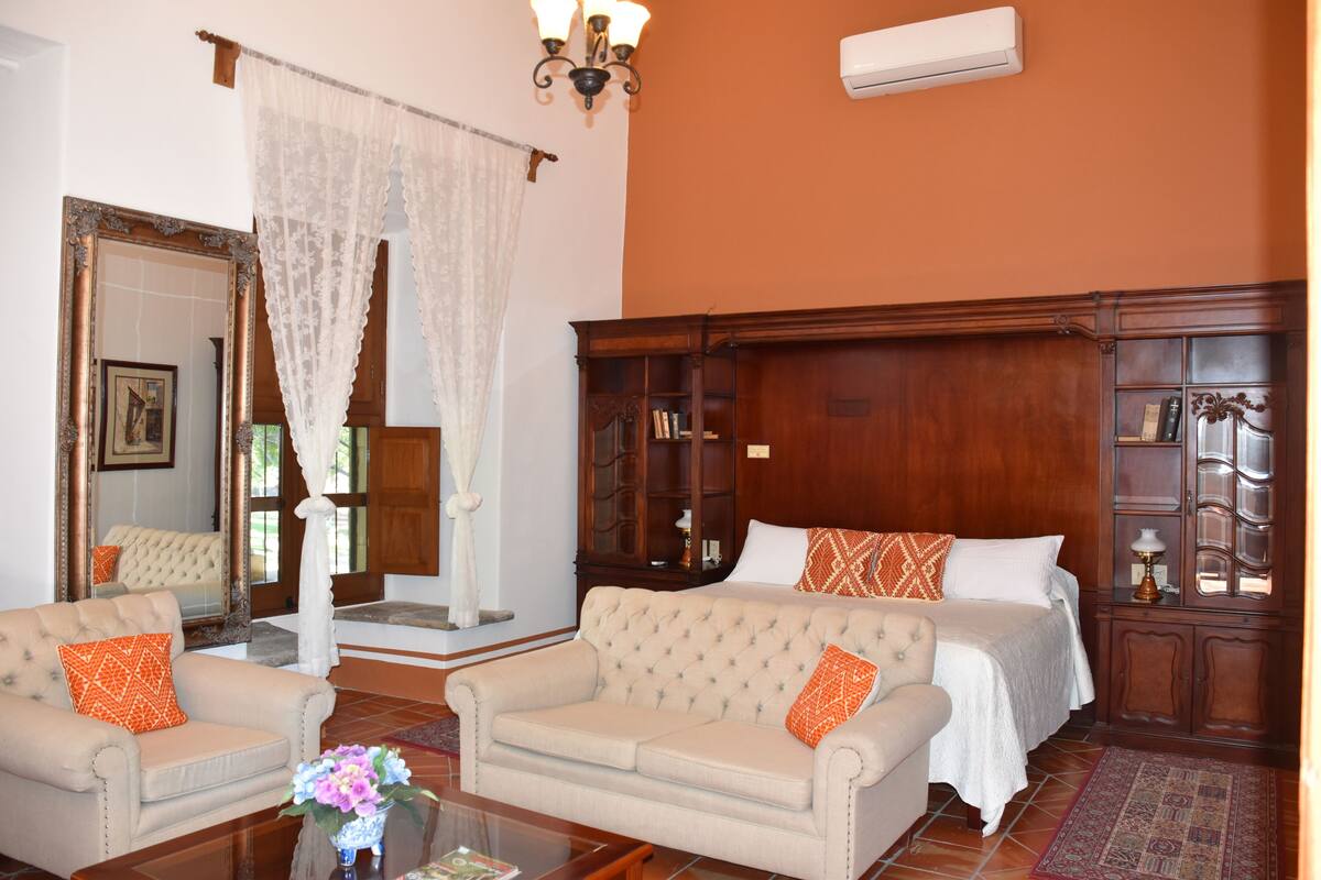 Una habitación de hotel con una cama grande, dos sillones, un armario de madera y un espejo.