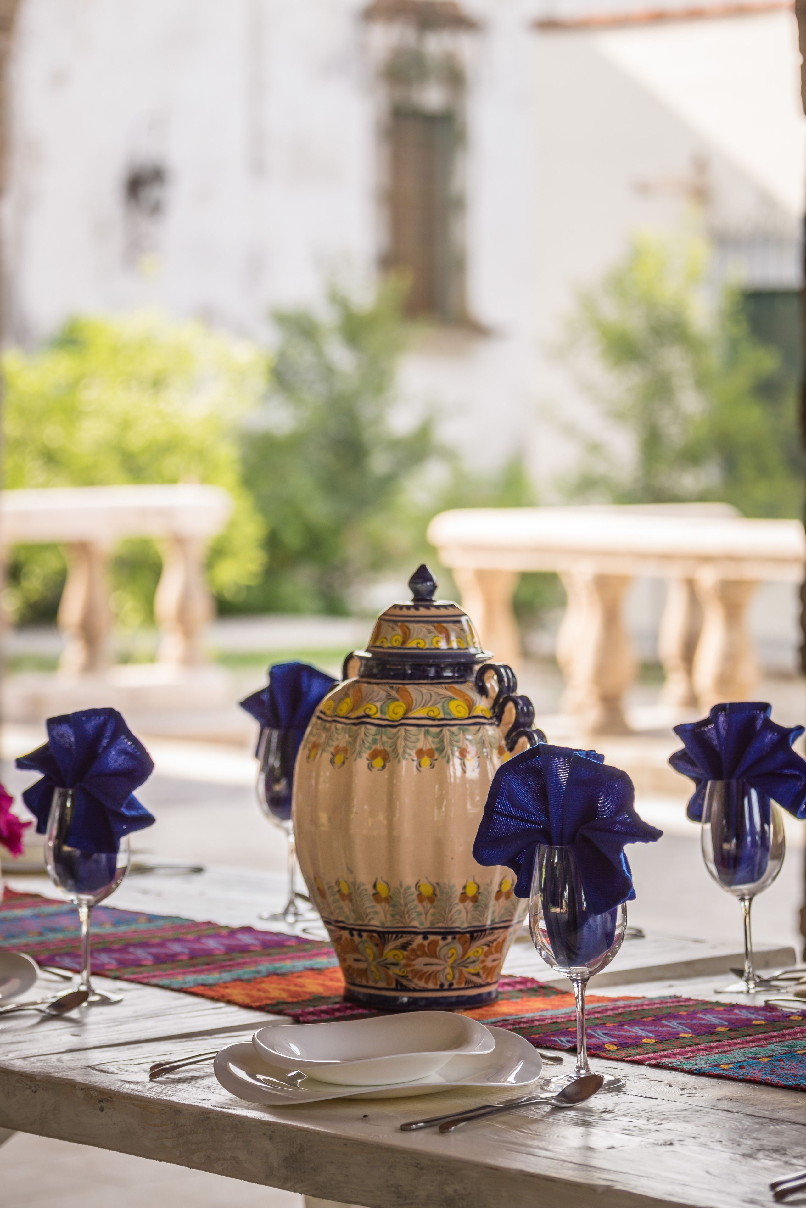 Una mesa dispuesta con una jarra de cerámica decorativa, copas de vino y un mantel individual colorido.