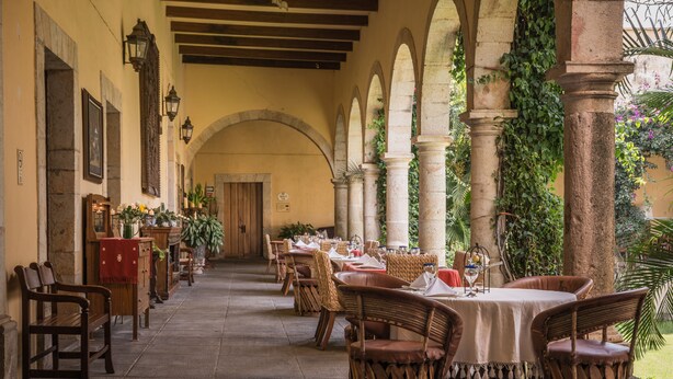 Un área de comedor al aire libre con mesas y sillas de mimbre, rodeada de columnas y arcos de piedra, con vista a un jardín.
