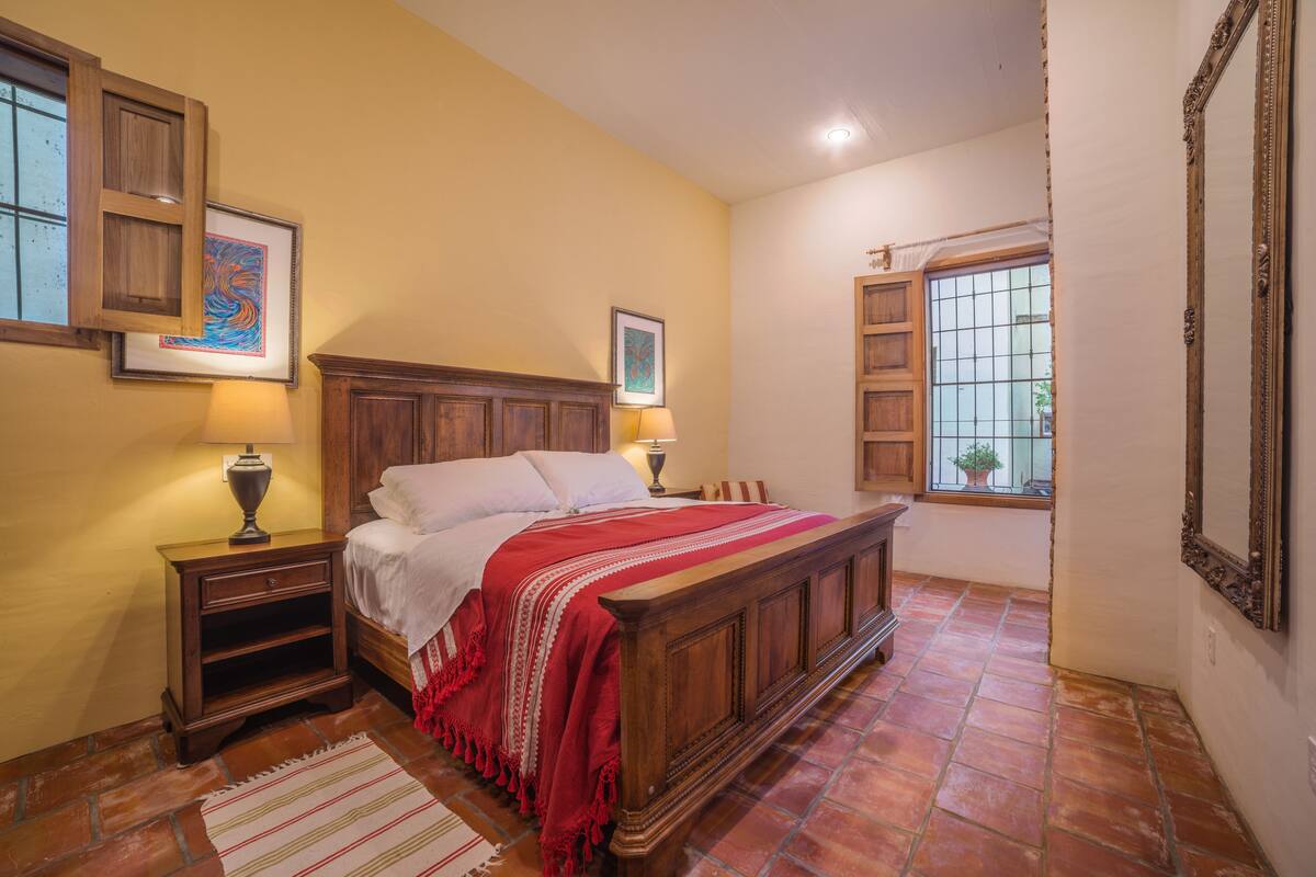 Un dormitorio con una cama de madera, dos mesitas de noche, una colcha roja, una ventana con vistas y un cuadro en la pared.