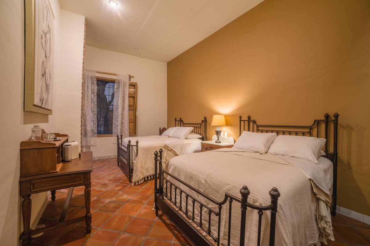 Un dormitorio con dos camas, una mesita de noche, una mesa y un ventanal con cortinas.