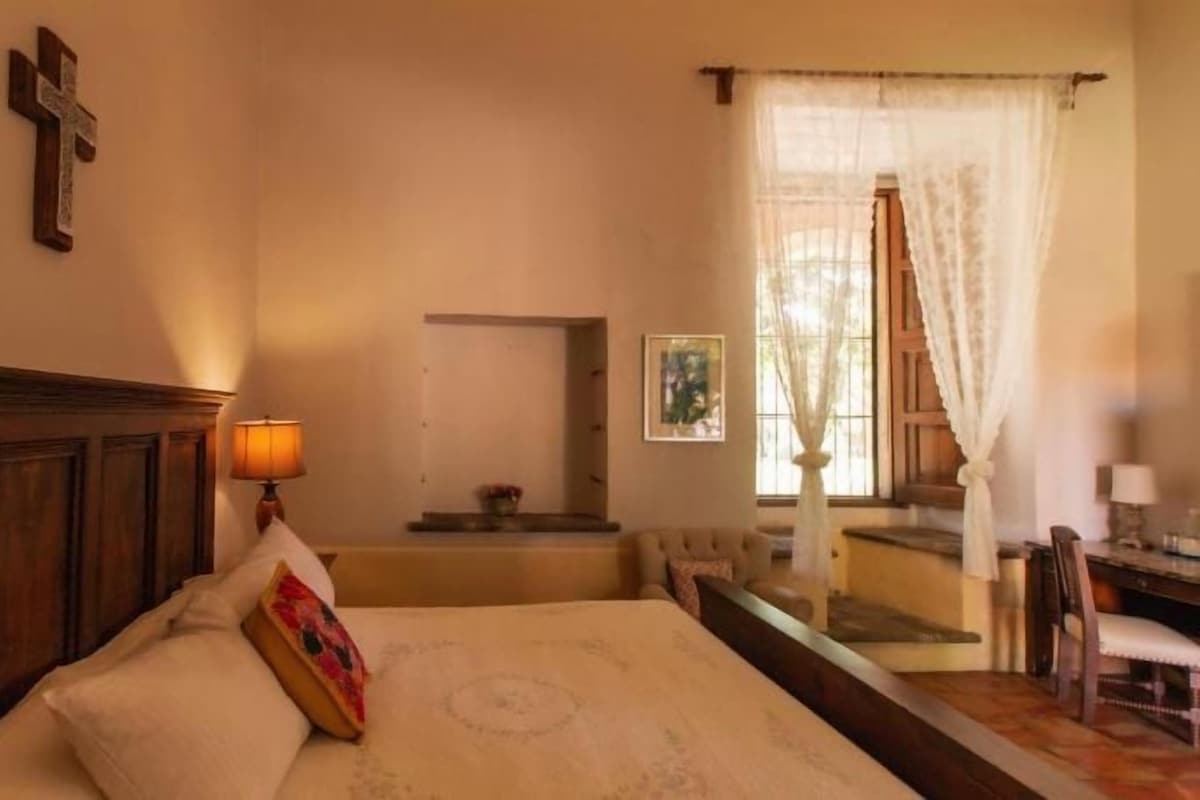 Une chambre à coucher avec une tête de lit en bois, une table de chevet avec une lampe, un lit recouvert d’une couvre-lit à motifs, une chaise et une fenêtre avec des rideaux.