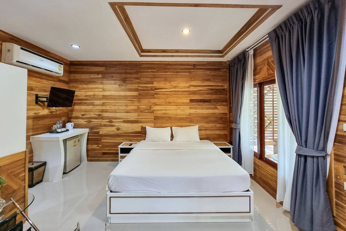 A bedroom with a wooden wall panel, a bed, a bedside table, a TV, and a window with curtains.