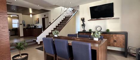 Cozinha moderna com ilha central, área de jantar com cadeiras azuis, escada e televisão de tela plana na parede.