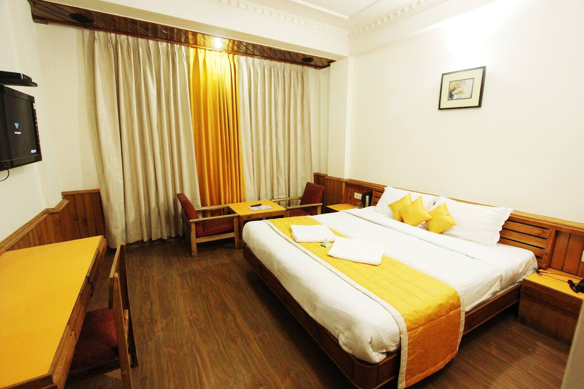 Habitación de hotel con cama, escritorio, silla, televisor y ventana con cortinas.