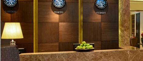 A reception desk with a wooden panel wall displaying clocks labeled 'LONDON', 'TOKYO', and 'SYDNEY'. A bowl of green apples is on the counter.