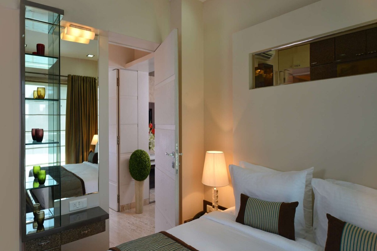 A hotel room with a large bed, a bedside table with a lamp, a mirror, and a glass cabinet with decorative items.