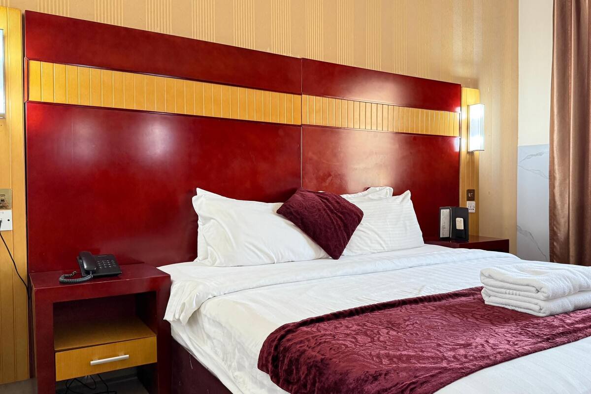 Habitación de hotel con cabecera roja, ropa de cama blanca, pared decorativa de mármol, mesita de noche de madera con un teléfono y una silla marrón.