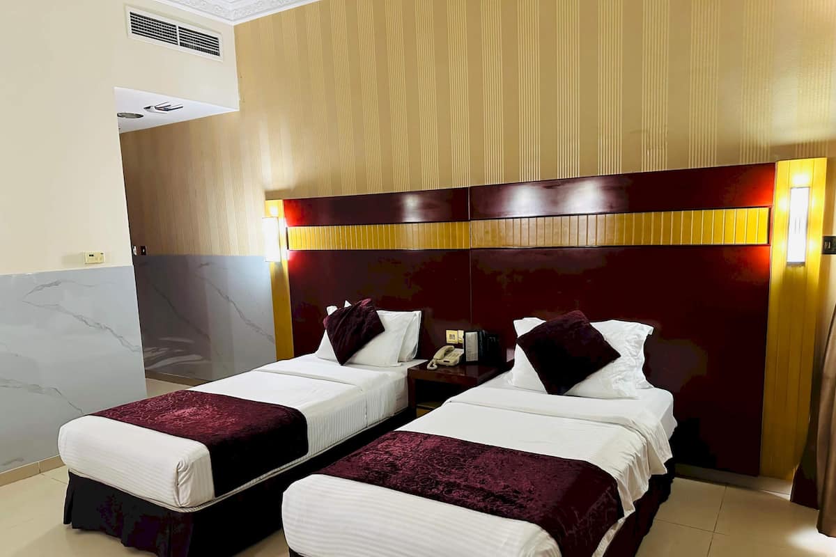 Habitación de hotel con dos camas individuales, cabecero de madera, mesita de noche con teléfono y rejilla de ventilación en la pared.