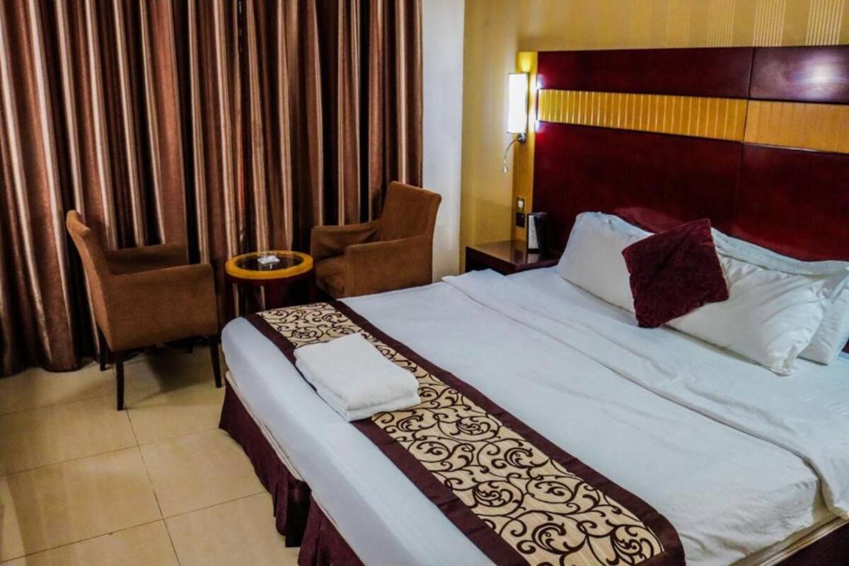 Una habitación de hotel con una cama grande, dos sillones, una mesita redonda y una mesita de noche con lámpara.