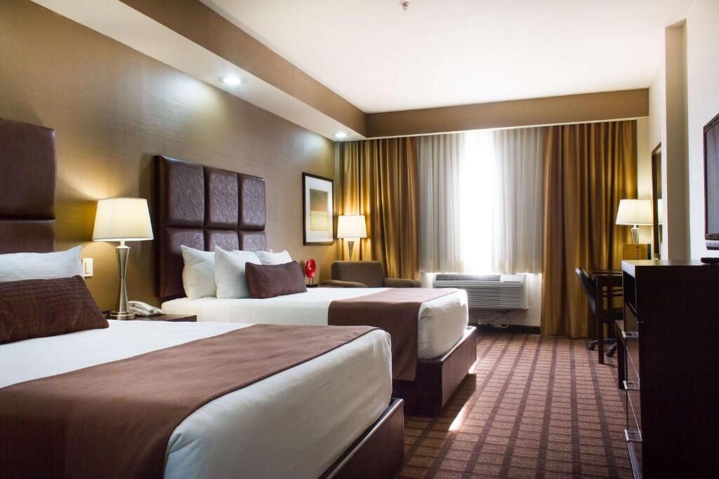 Ein Hotelzimmer mit zwei Betten, einem Schreibtisch, einem Stuhl, einem Fernseher und einem großen Fenster mit Vorhängen.