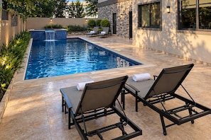 Una piscina rectangular con cascada, rodeada por un muro de piedra y un patio con sillones y una mesa.