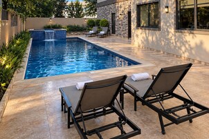Una piscina rectangular con cascada, rodeada por un muro de piedra y un patio con sillones y una mesa.