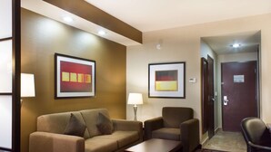 Un vestíbulo de hotel con un sofá, un sillón y una mesita. Hay dos obras de arte enmarcadas en la pared, una puerta y una lámpara.