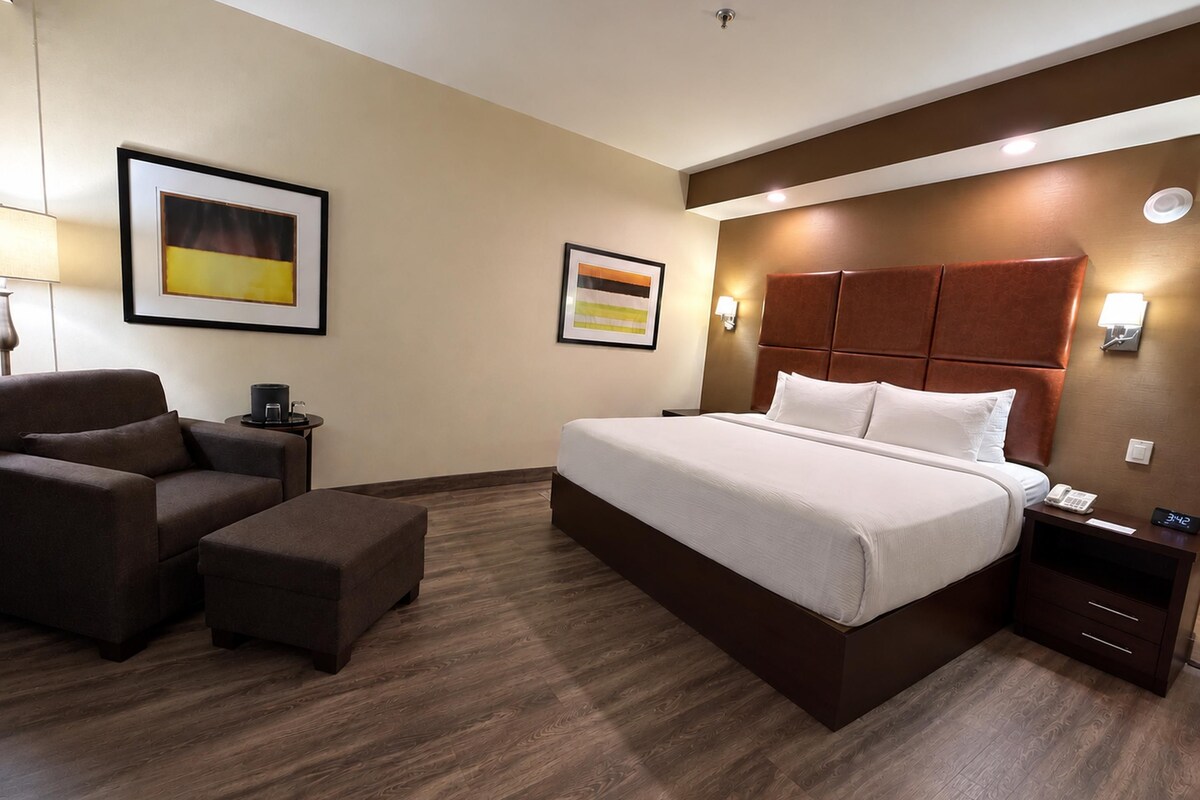 Una habitación de hotel con una cama grande, una silla, una mesita pequeña y dos lámparas de pared.