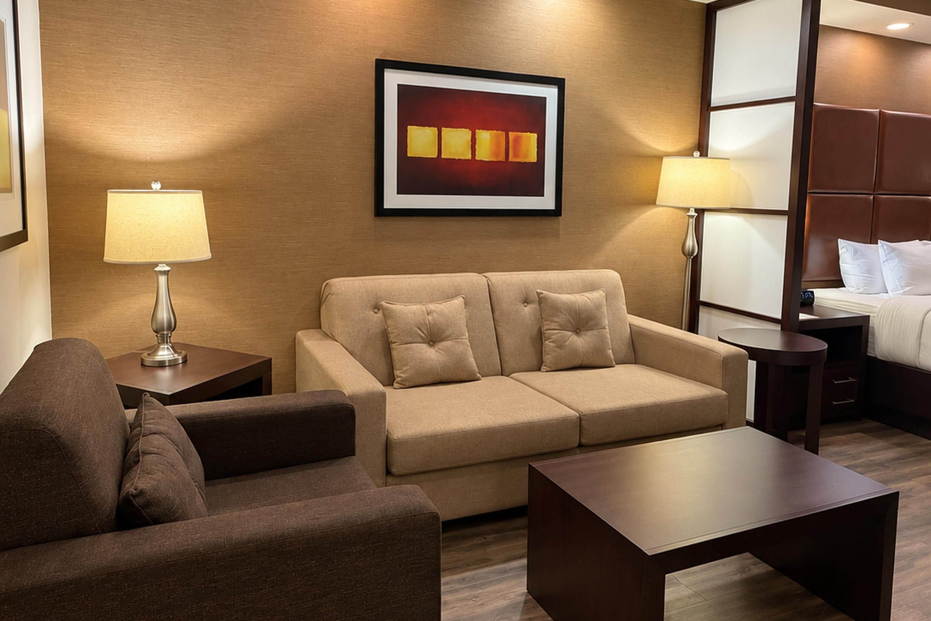 Habitación de hotel con sofá, mesa de centro, cama con cabecera, mesita de noche, lámpara y un cuadro en la pared.