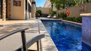 Una piscina con borde de azulejos, un área de descanso con sillas y un edificio de fachada de piedra.