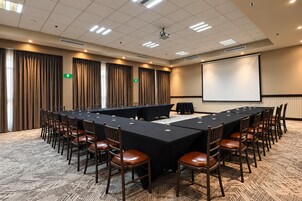 Un salón de conferencias con una larga mesa, sillas, una pantalla de proyector y cortinas.
