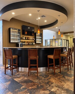 Zona de bar con una barra curva, varios taburetes, un televisor montado sobre la barra, y una variedad de botellas y vasos en estanterías.
