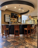 Zona de bar con una barra curva, varios taburetes, un televisor montado sobre la barra, y una variedad de botellas y vasos en estanterías.