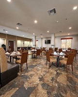 Um espaço de refeições de hotel, com várias mesas e cadeiras, uma televisão na parede e uma área de cozinha ao fundo.