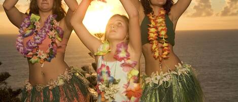 Three individuals in traditional attire with leis, standing by a body of water during sunset.