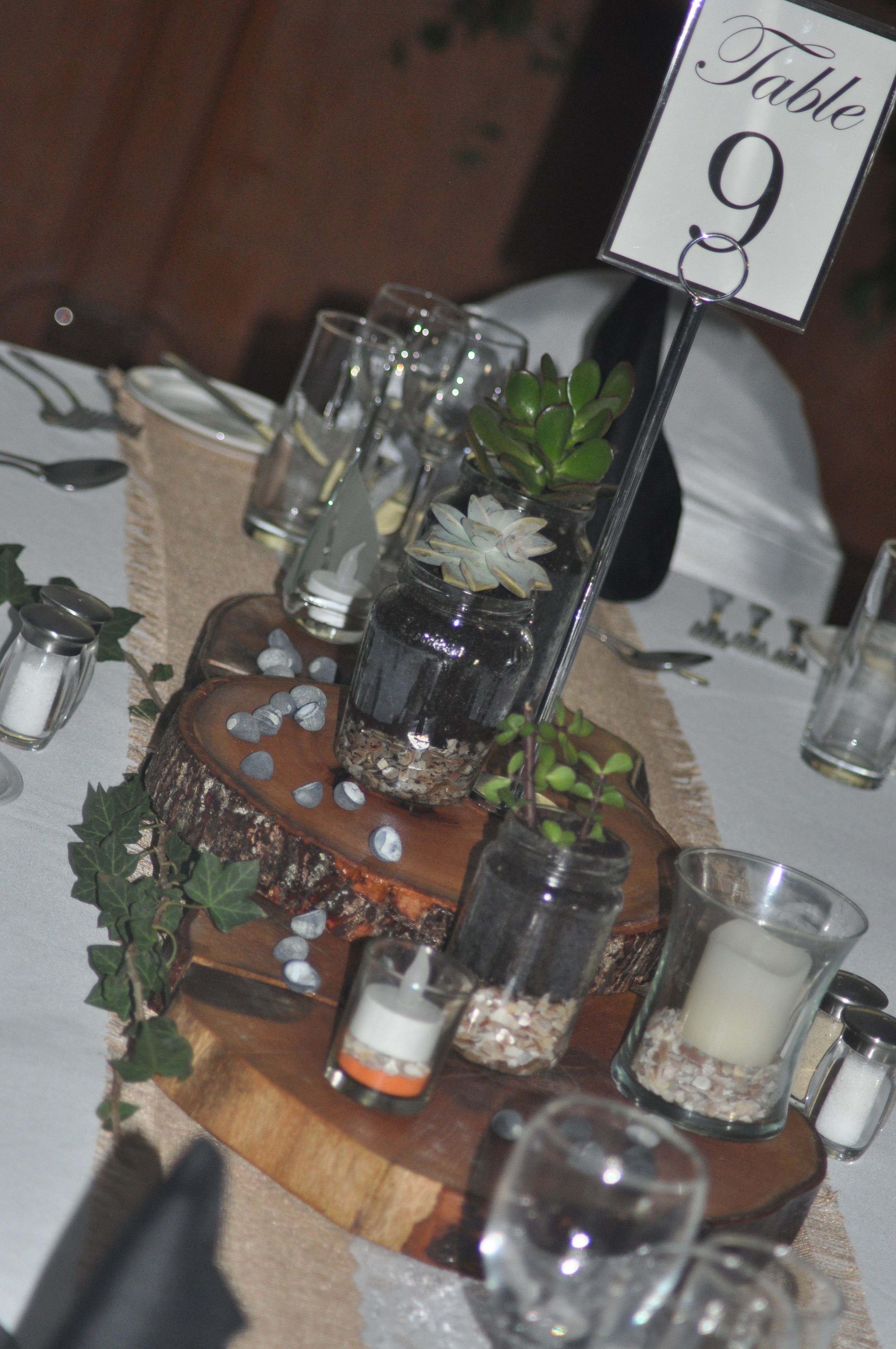 Una mesa dispuesta con frascos de vidrio que contienen plantas y velas, un centro de mesa de madera y una señal con el número de la mesa.