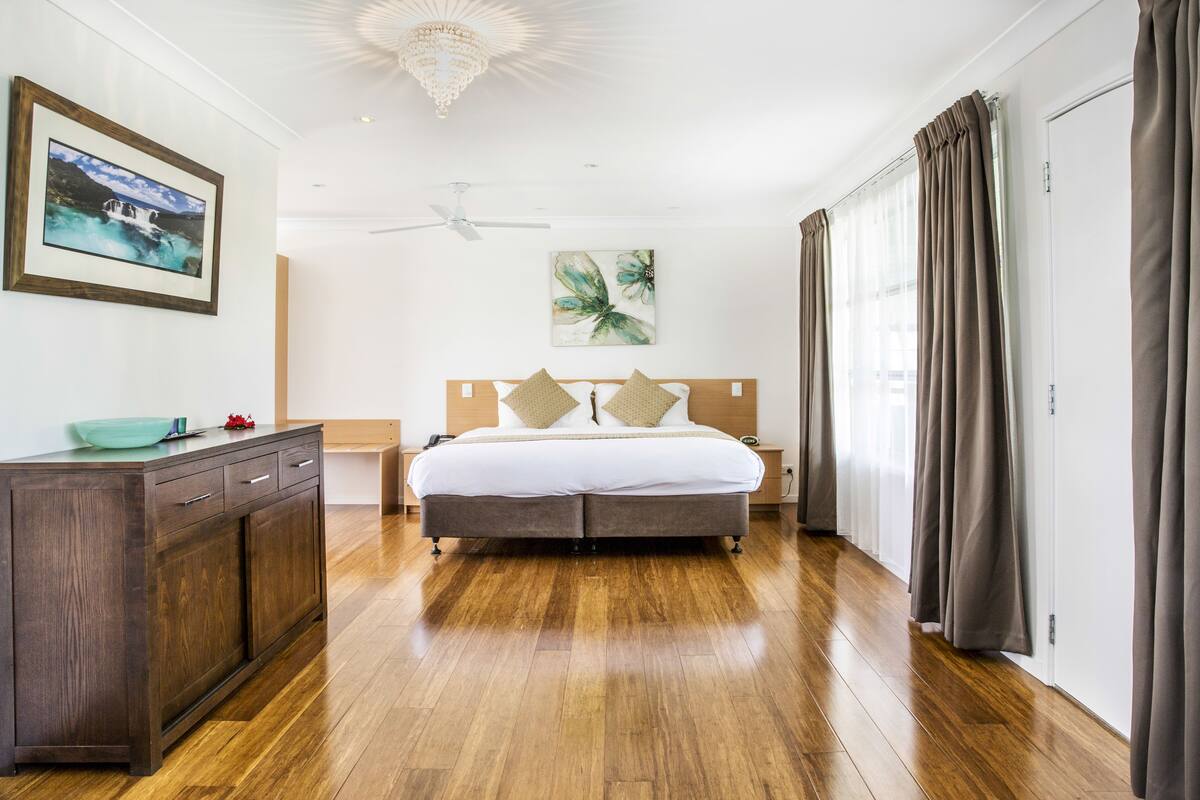 A bedroom with a wooden bed, a nightstand, a dresser, a ceiling fan, and a large window with curtains.
