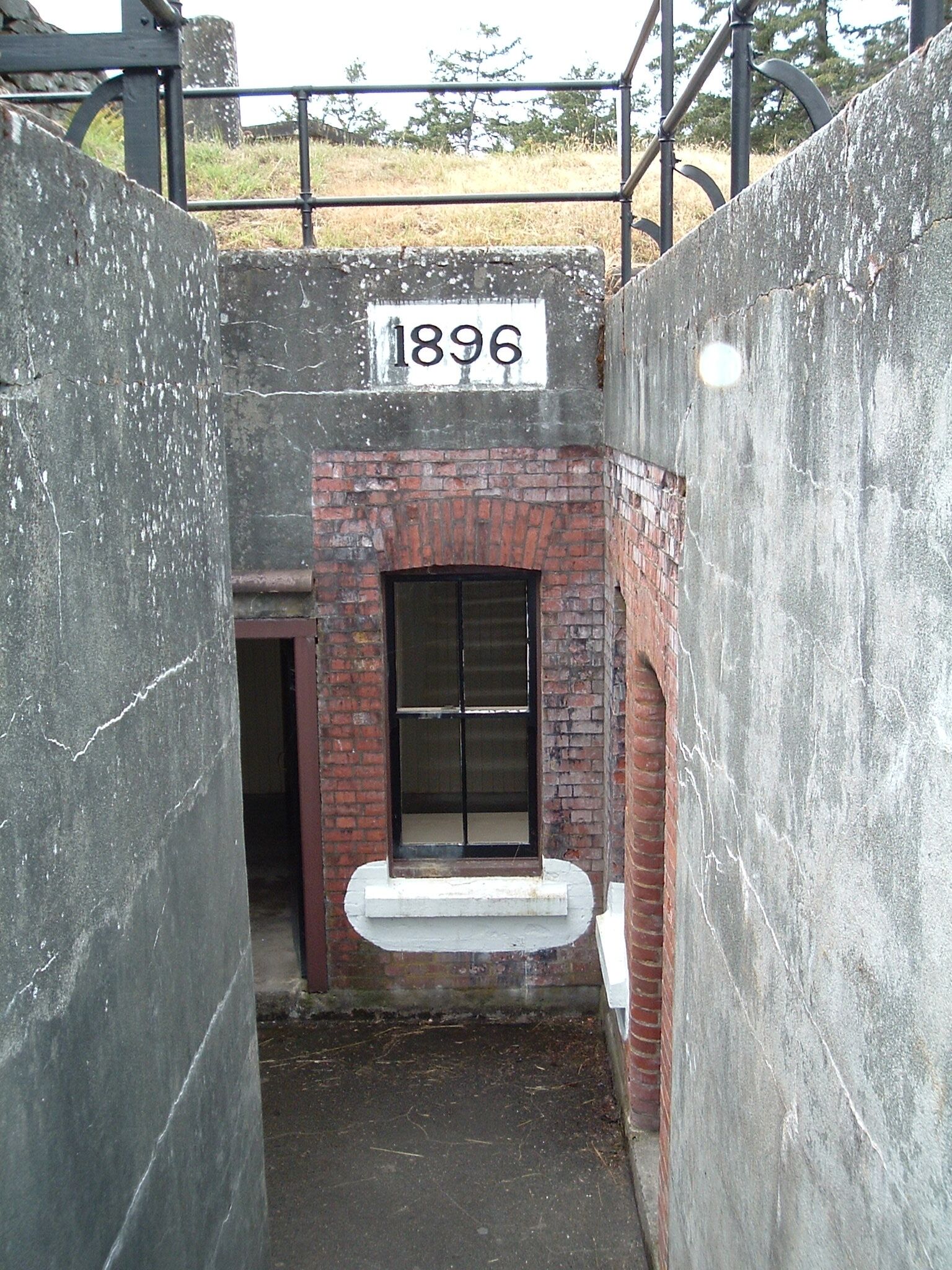 A narrow, enclosed space with a brick wall and a metal gate. A sign with the number "1896" is visible.