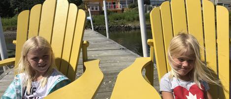 Two children sitting on yellow Adirondack chairs on a wooden dock with a house in the background.