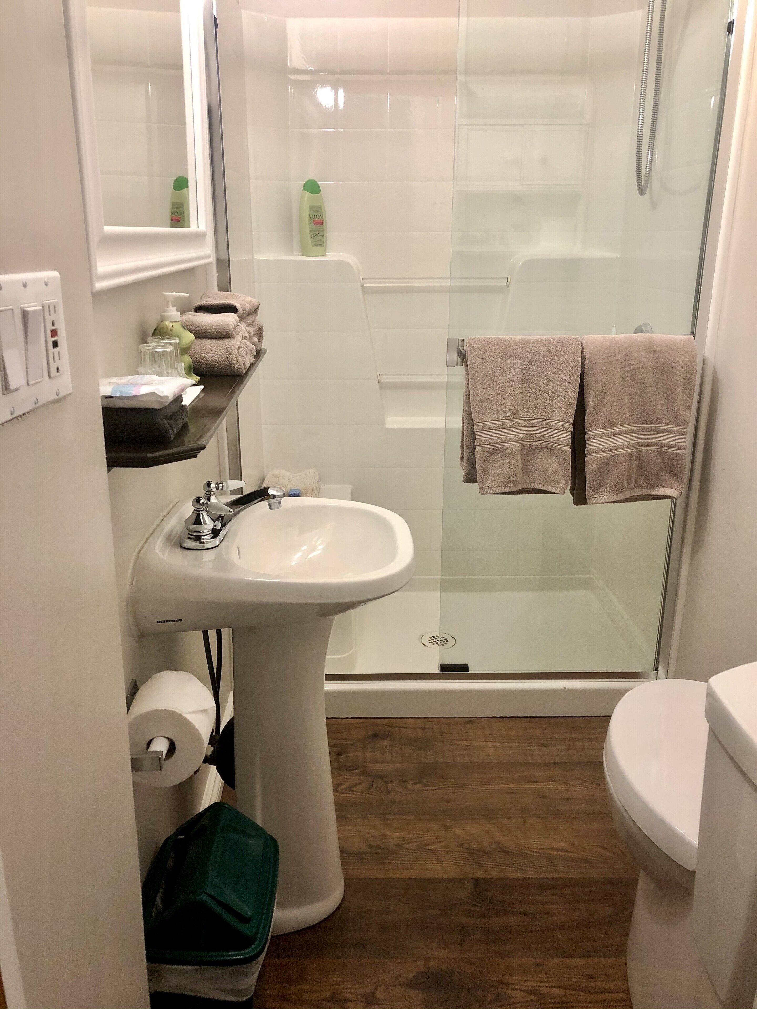 A bathroom with a glass-enclosed shower, a pedestal sink, and a toilet. There are towels on a shelf and a green trash bin.