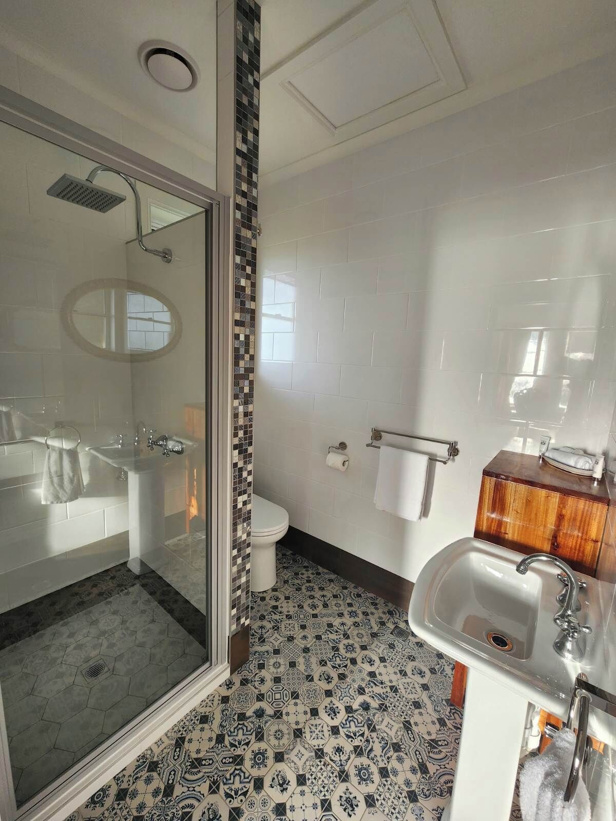 Un baño con cabina de ducha de vidrio, inodoro blanco, lavamanos con tocador de madera y baldosas de suelo estampadas.