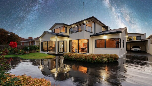 Una casa moderna de dos pisos con ventanas grandes y un balcón, rodeada de un jardín y un estanque reflectante.
