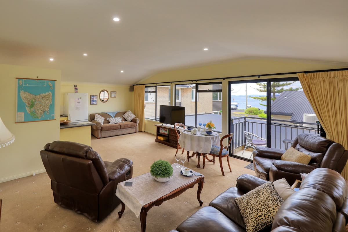 Una sala de estar con sofá, sillón, mesa de centro y televisor. Hay un balcón con vista al mar.
