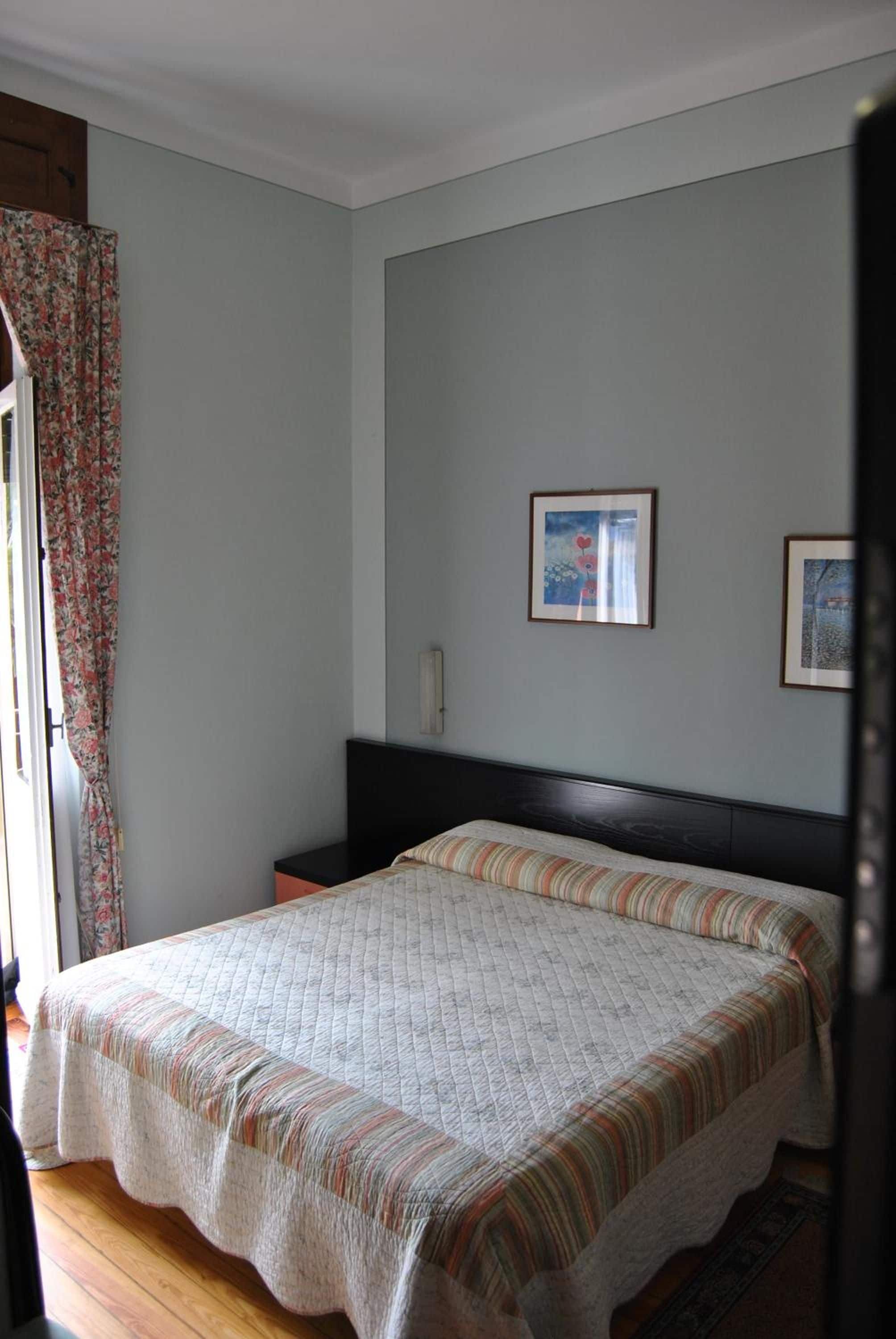 Un dormitorio con cama, mesita de noche, ventana con cortinas y dos cuadros en la pared.