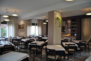 A restaurant with multiple tables and chairs, a bar area with a wine rack, and a large painting on the wall. - Hotel Villa Mon Toc (Stresa)