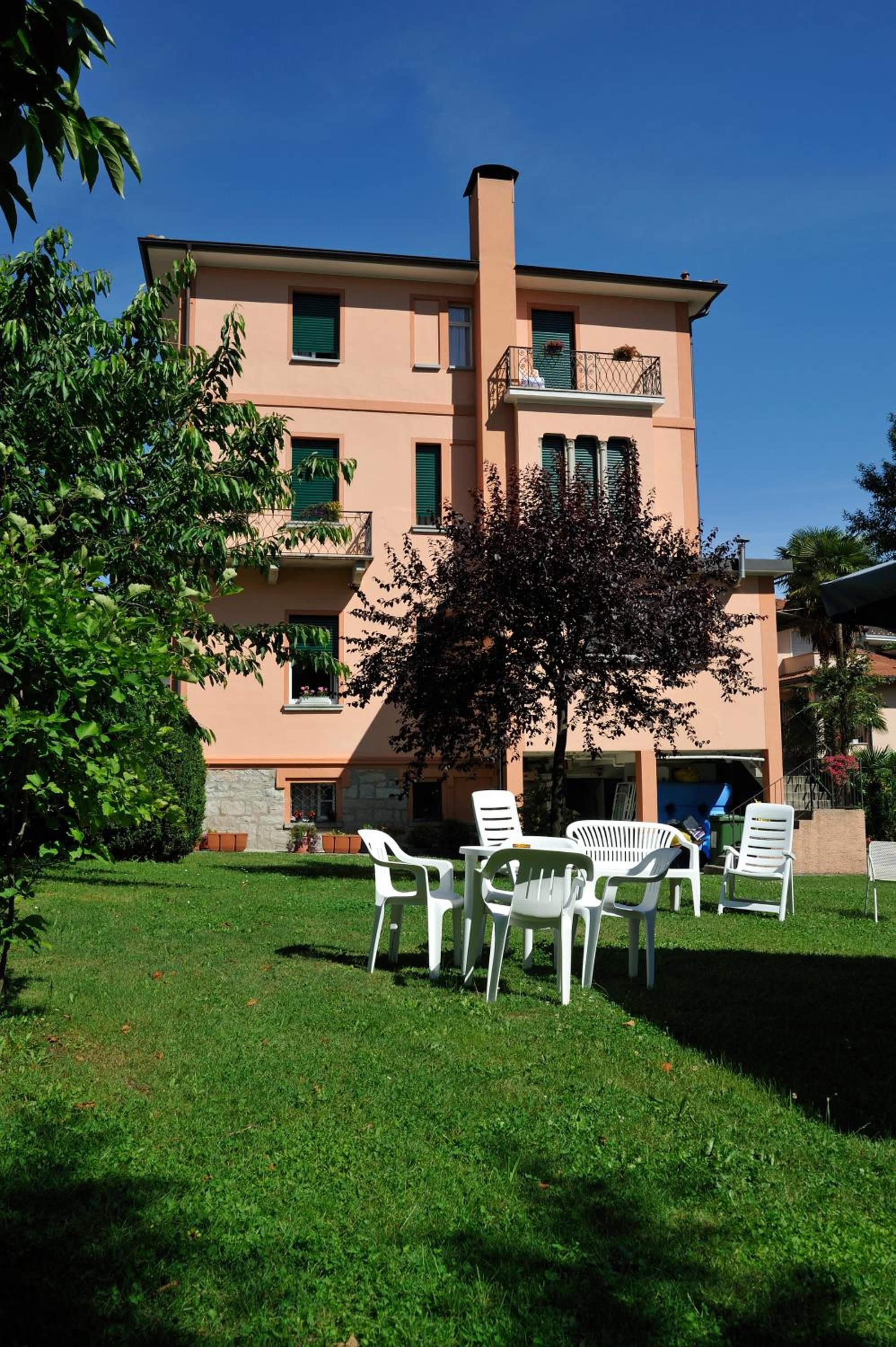 Un bâtiment à deux étages, doté d’un toit plat, d’un balcon avec une balustrade et d’une cheminée. On y voit une pelouse avec plusieurs chaises en plastique blanc et un arbre au premier plan.