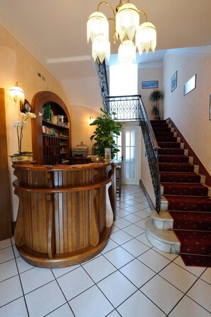 A reception area with a wooden counter, a staircase, and a chandelier. - Hotel Villa Mon Toc (Stresa)