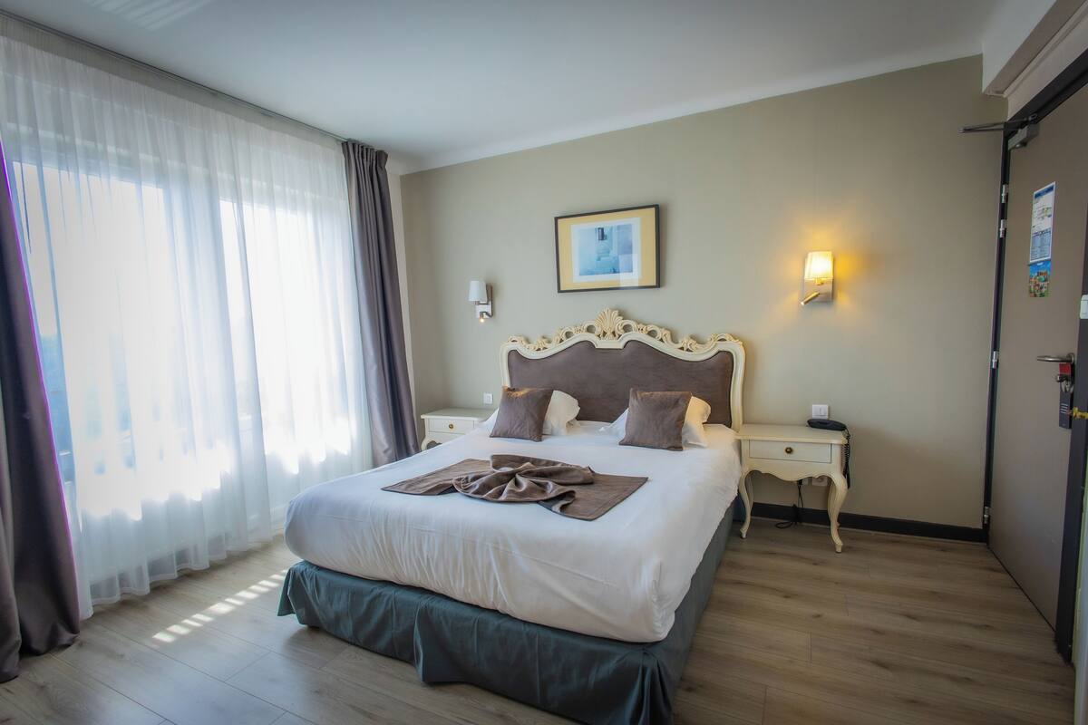 Habitación de hotel con una cama grande, una mesita de noche, una lámpara de pared y un ventanal con cortinas.