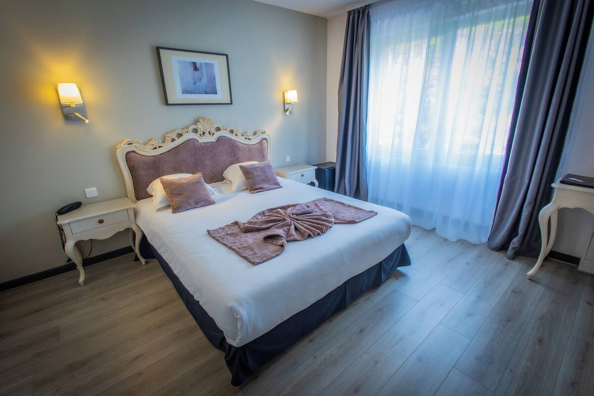 Una cama bien hecha con un cabecero decorativo, dos mesitas de noche y un amplio ventanal con cortinas.