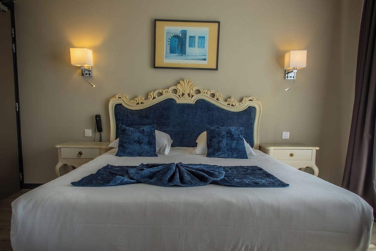 Una cama bien hecha con una decoración en blanco y azul, dos mesitas de noche con lámparas, un cuadro en la pared y suelo de madera.