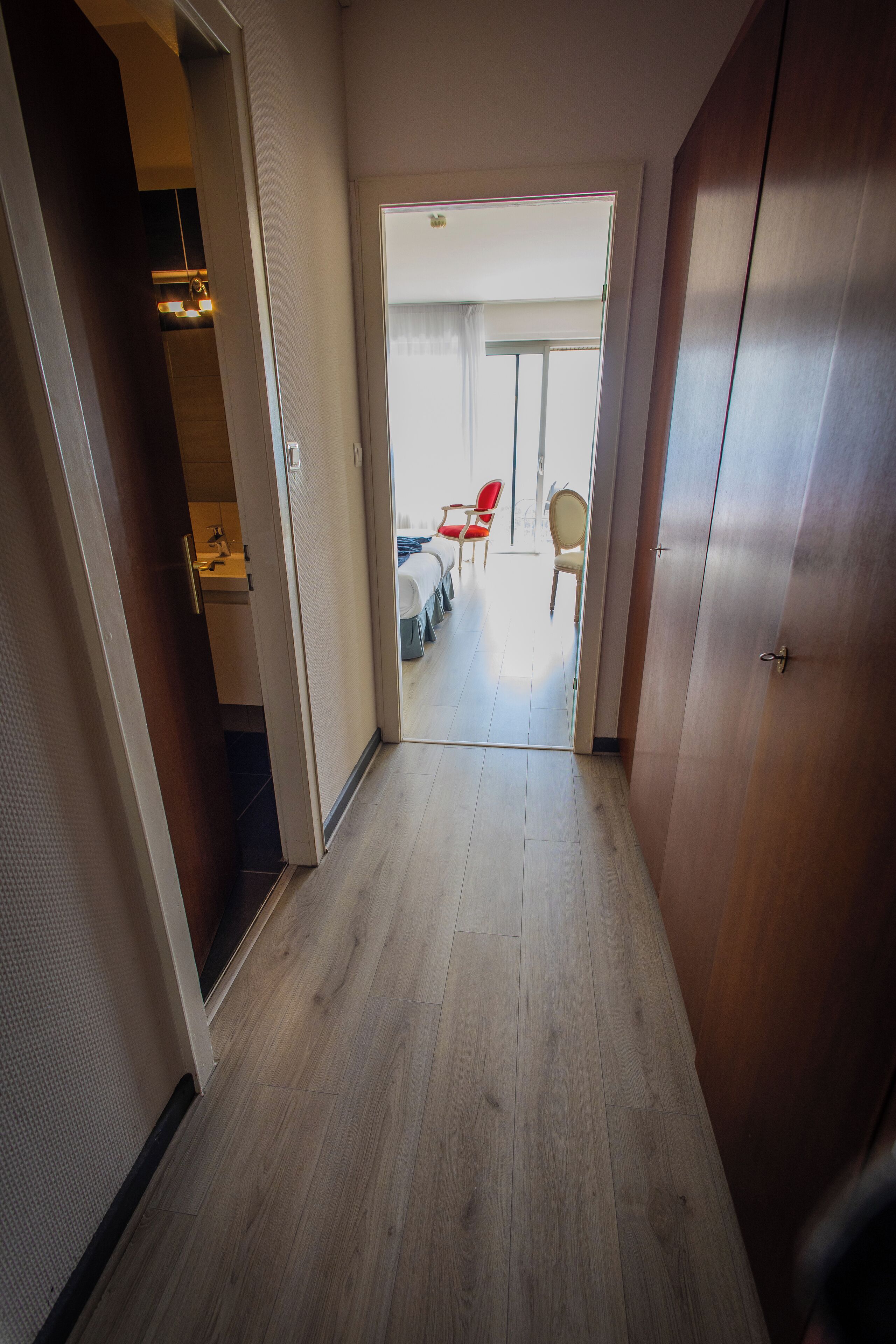 Un couloir au plancher de bois qui mène à une pièce contenant un lit et des chaises.