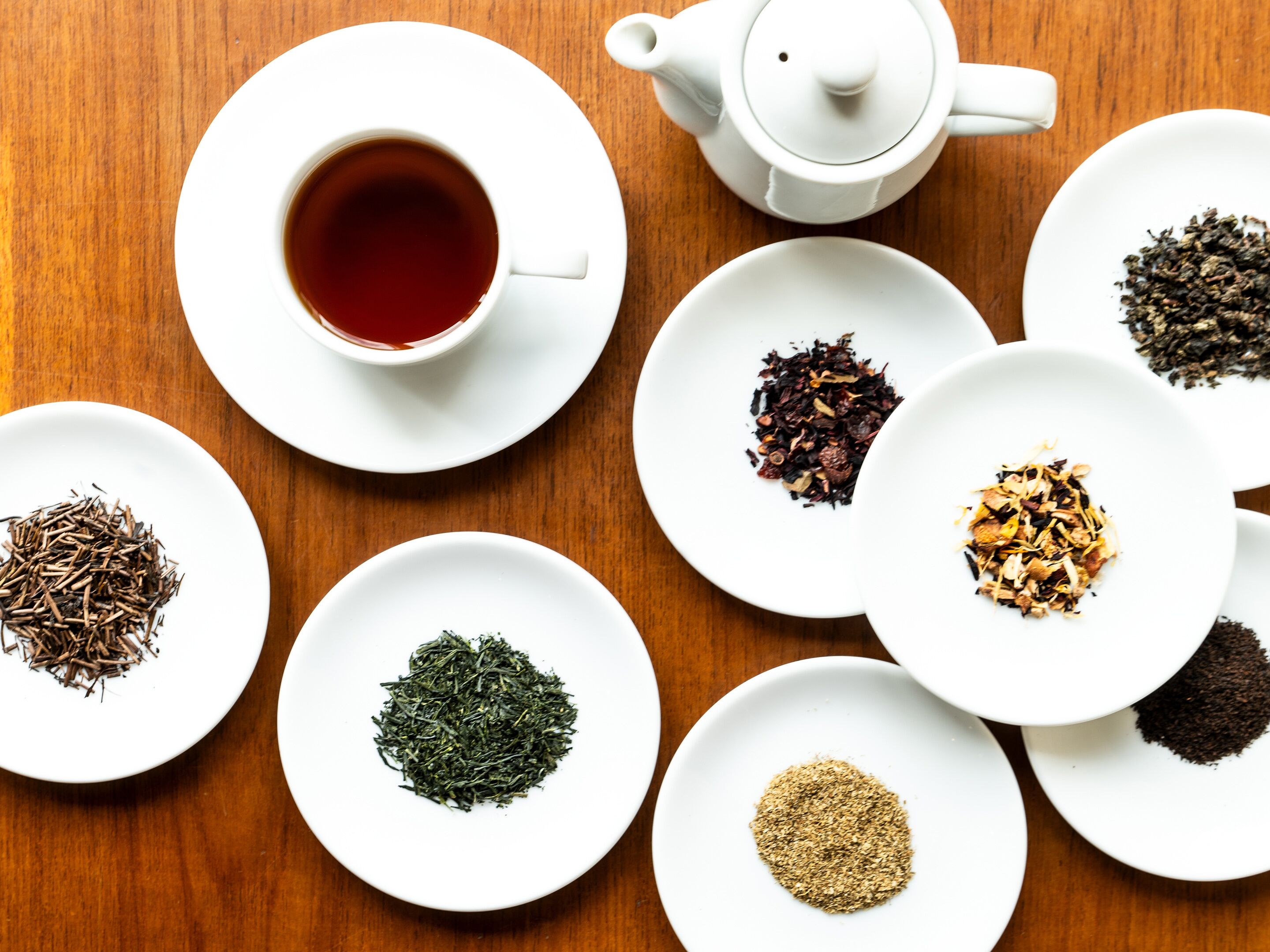 Variedad de hojas de té sobre platos blancos dispuestos en una mesa de madera, con una tetera y una taza de té a la vista.