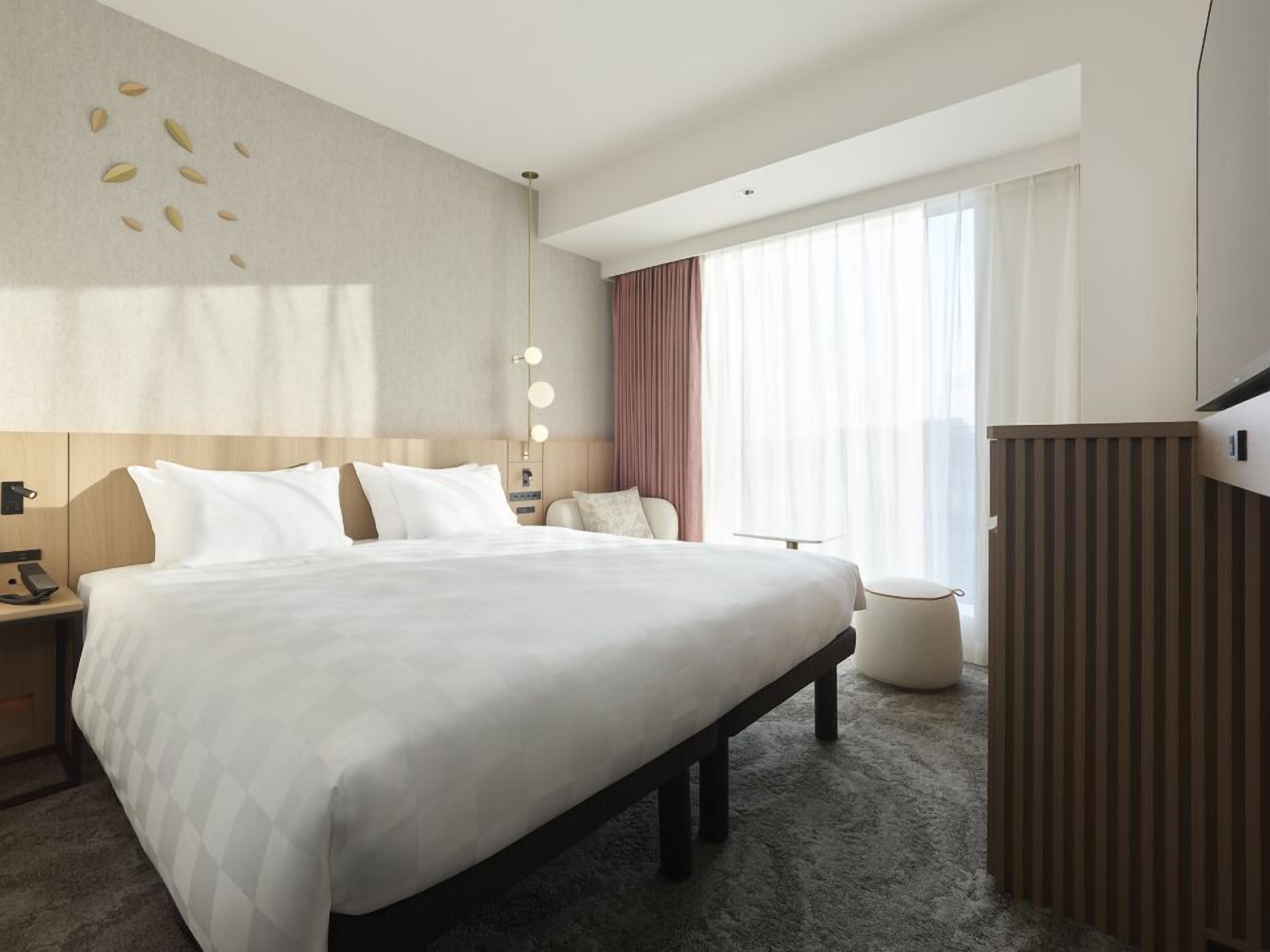 Una habitación de hotel con una cama grande, mesitas de noche, un escritorio, una silla, un otoman redondo, un armario y un ventanal con cortinas.