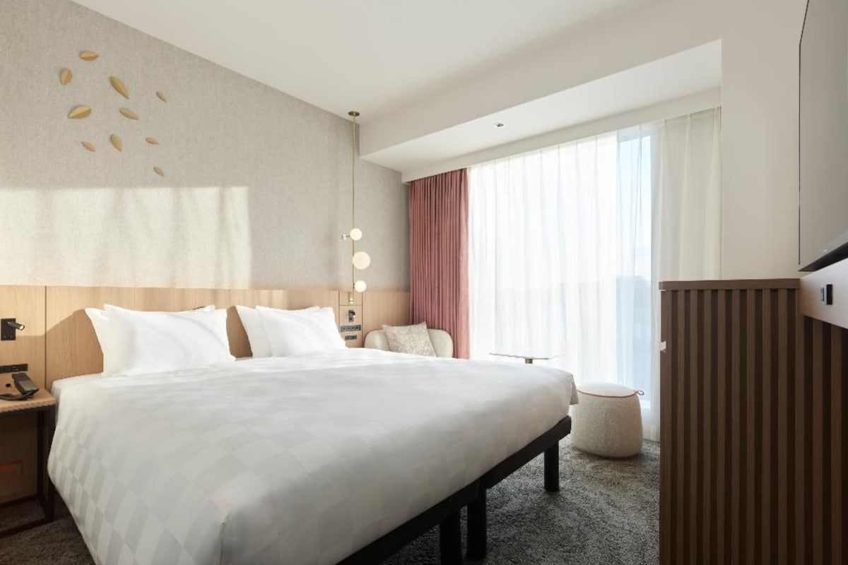Habitación de hotel con una cama grande, mesitas de noche, un escritorio, una silla, un otoman redondo, un armario y un ventanal con cortinas.