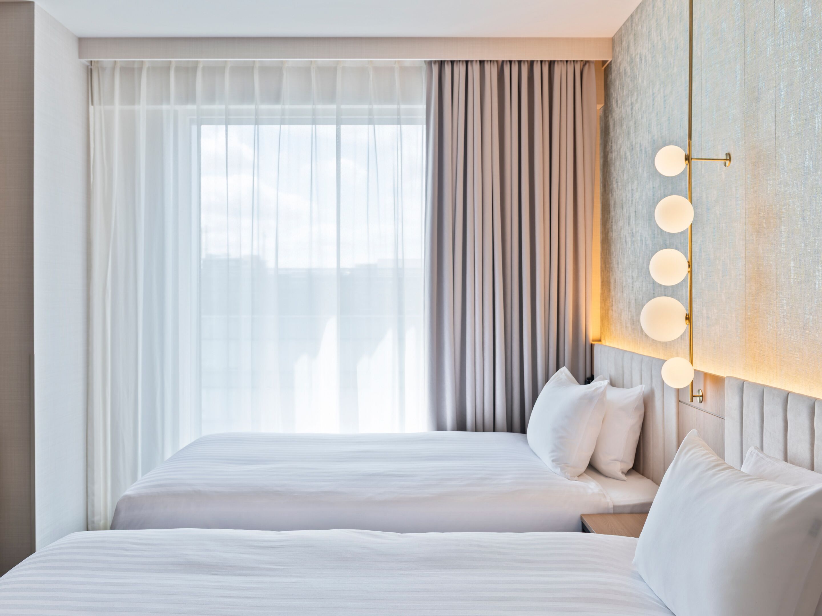 Une chambre d’hôtel avec deux lits, une grande fenêtre avec des rideaux légers, un luminaire fixé au mur et un mur à texture.